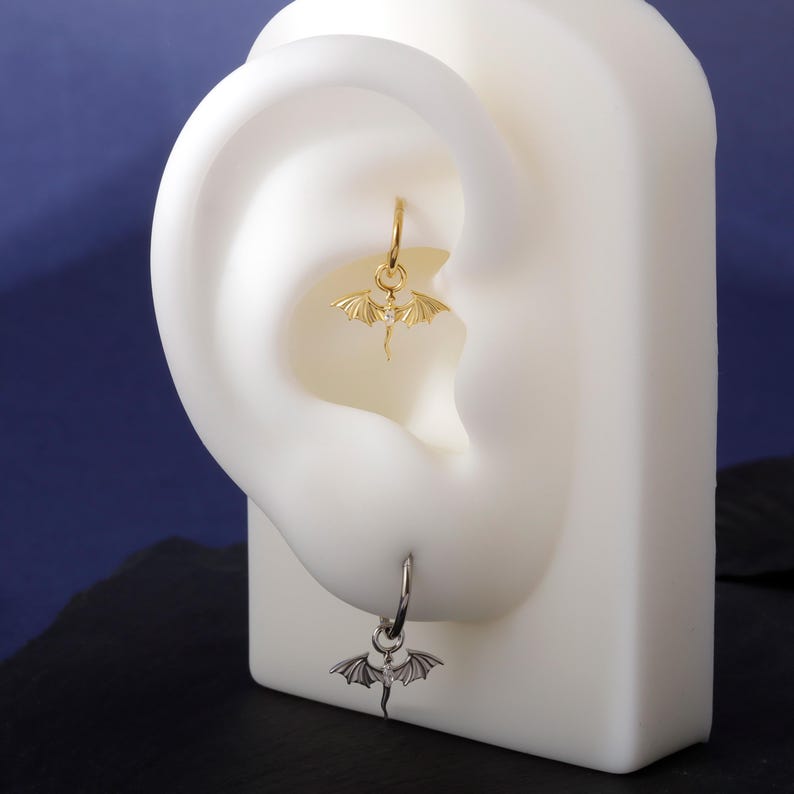 May include: A pair of dragon earrings, one gold and one silver. The gold earring has a dragon pendant with a gemstone, suspended from a hoop. The silver earring also features a dragon pendant. Both are shown on a white ear model against a dark blue backdrop.