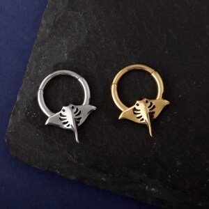 May include: Two hoop earrings, one silver and one gold, each featuring a stylized manta ray charm. The earrings are displayed on a dark, textured surface against a dark blue background. The manta ray charms have detailed cut-out designs.