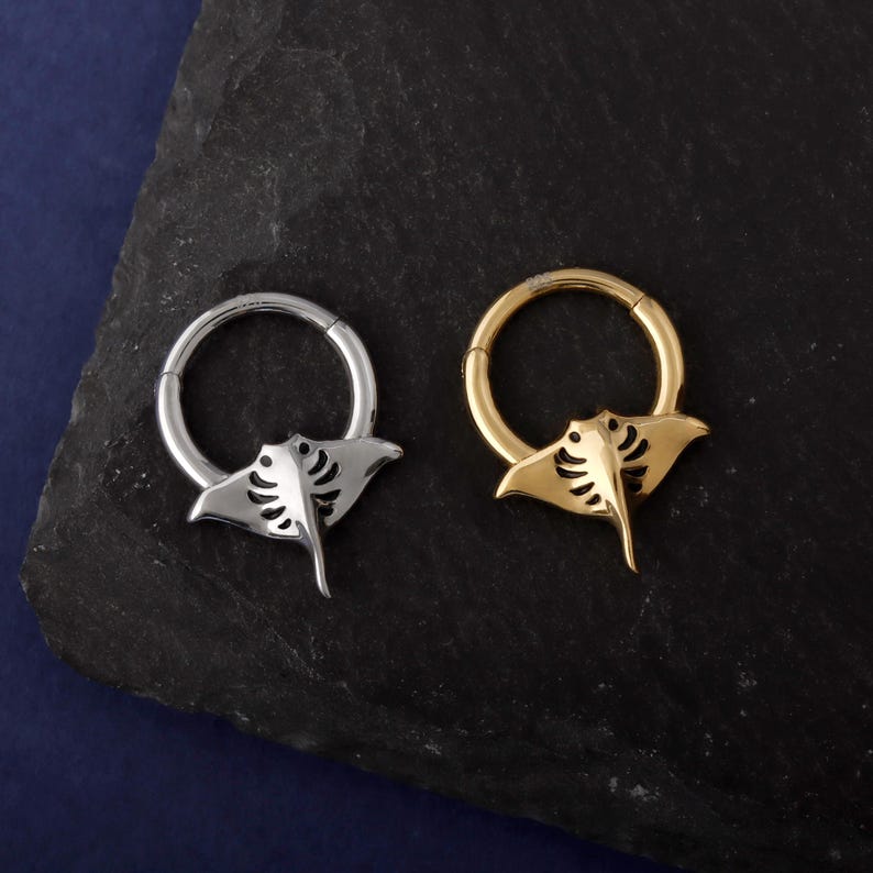 May include: Two hoop earrings, one silver and one gold, each featuring a stylized manta ray charm. The earrings are displayed on a dark, textured surface against a dark blue background. The manta ray charms have detailed cut-out designs.