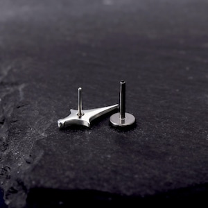 May include: A silver-toned metal body jewellery piece featuring a flat-backed post and a decorative front element shaped like a four-pointed star or dagger. The item has a polished, reflective surface and a straight, thin post extending from the back.