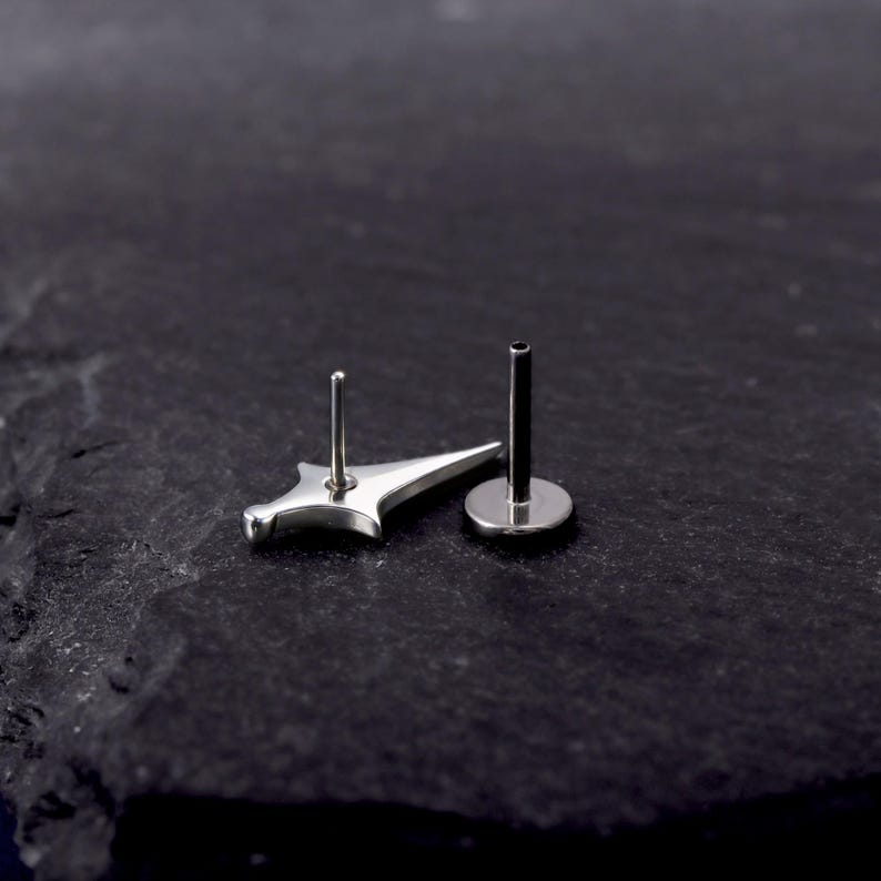 May include: A silver-toned metal body jewellery piece featuring a flat-backed post and a decorative front element shaped like a four-pointed star or dagger. The item has a polished, reflective surface and a straight, thin post extending from the back.
