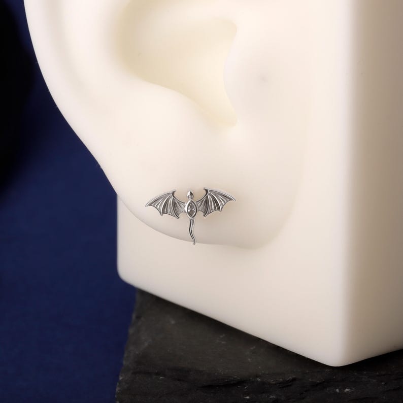 May include: A silver dragon stud earring with detailed wings and a slender tail. The earring is shown on a white ear model against a dark blue background. The dragon's body features a small, oval-shaped gem.