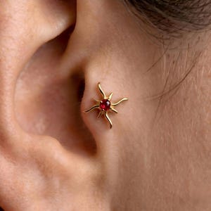 May include: A gold ear piercing stud featuring a central round red gemstone surrounded by four curved, pointed rays extending outward in a starburst pattern. The jewelry is positioned in the tragus area of the ear, showcasing a polished metal finish.