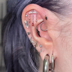May include: An ear with multiple piercings featuring silver-toned jewelry, including a bat-shaped charm with hanging chains and a red teardrop gem, a baguette-cut stone, a sword-shaped stud, a black crescent moon, and various small metallic hoops.