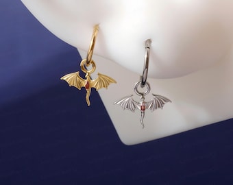 May include: Two hoop earrings featuring a dangling bat-shaped charm with a small red gemstone set in the centre. One earring is gold-toned, and the other is silver-toned. Both charms display detailed wings and a pointed tail design.