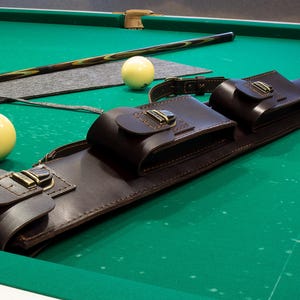 May include: A brown leather pool cue case with three compartments, secured by brass-colored buckles, rests on a green pool table. Two ivory-colored billiard balls and a pool cue are also visible.