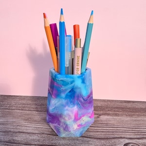 May include: A blue, purple, and white marbled resin pen holder filled with colorful pencils and pens. The pen holder has a geometric design and is sitting on a wooden surface. The background is a solid pink color.