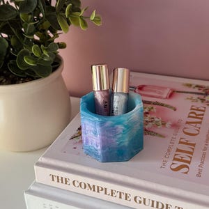May include: A hexagonal resin holder in shades of blue, purple, and white, holding two small bottles with silver caps. The holder is on a stack of books titled "The Complete Guide to Self Care" and "The Complete Guide to SE". A potted plant is visible.