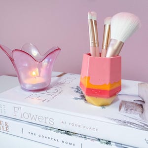 May include: A pink and orange geometric makeup brush holder with three brushes and a large fluffy brush. A pink tulip-shaped candle holder with a lit candle sits next to it. The items are on a book.
