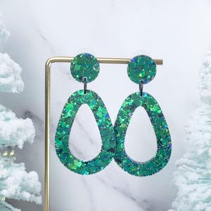 May include: Green glitter teardrop earrings. These dangle earrings feature a circular stud and a large, open teardrop shape. The earrings are a vibrant green color and are displayed on a gold earring stand. The background includes a marble surface and a frosted Christmas tree.