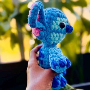 May include: A handmade blue crochet character with pink detailing on the ears. The character has a dark blue nose and black eyes. The text "@AmayezingCrafts" is visible at the bottom of the image. The plush toy is held in a hand.