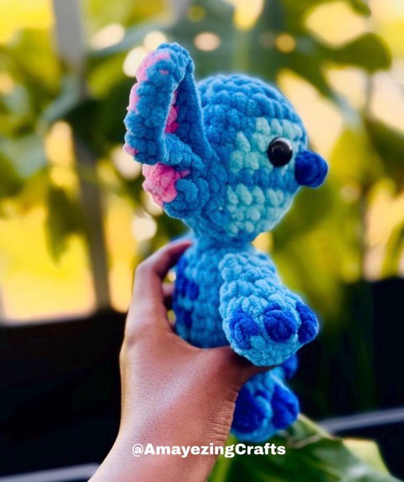May include: A handmade blue crochet character with pink detailing on the ears. The character has a dark blue nose and black eyes. The text "@AmayezingCrafts" is visible at the bottom of the image. The plush toy is held in a hand.