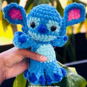 May include: A handmade blue crochet plush toy with large ears, black eyes, and a light blue belly. The plush has a textured appearance and is held in a hand. The text "@AmayezingCrafts" is visible at the bottom of the image.
