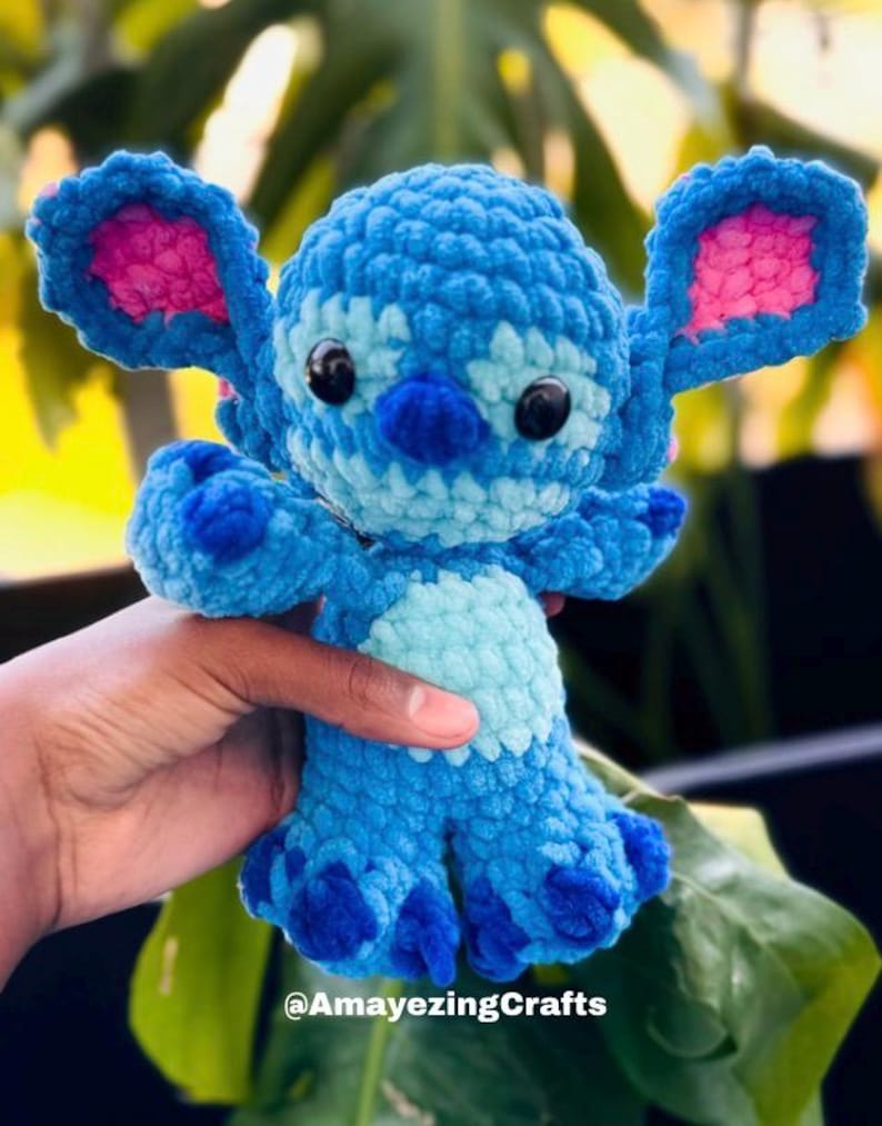 May include: A handmade blue crochet plush toy with large ears, black eyes, and a light blue belly. The plush has a textured appearance and is held in a hand. The text "@AmayezingCrafts" is visible at the bottom of the image.