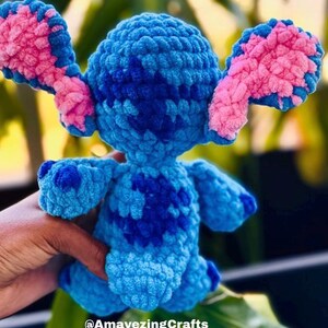 May include: A handmade blue and pink crochet Stitch doll. The plush toy features large ears with pink accents, a dark blue pattern on the body, and is held by a hand. The text "@AmayezingCrafts" is visible.