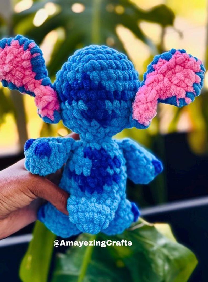 May include: A handmade blue and pink crochet Stitch doll. The plush toy features large ears with pink accents, a dark blue pattern on the body, and is held by a hand. The text "@AmayezingCrafts" is visible.