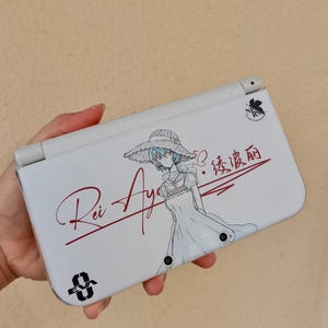 May include: A white handheld gaming console featuring a graphic of a character wearing a wide-brimmed hat. The text "Rei Ayanami" is written in red script, alongside Japanese characters, an "EVA 0" logo, and a small "NERV" emblem in the corner.