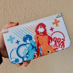 Puede incluir: Una consola de juegos portátil blanca con un estampado gráfico de dos personajes de anime con cabello azul y rojo. El diseño incluye signos más azules y naranjas, patrones circulares y el texto 002 EVA LANGLEY impreso en rojo a la derecha.