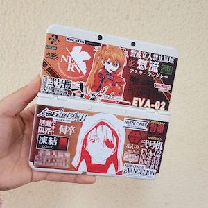 May include: A white handheld gaming console featuring custom decals from Neon Genesis Evangelion. The top screen area displays Asuka Langley, while the bottom area features a character with an eyepatch, Japanese text, and EVA-02 branding.