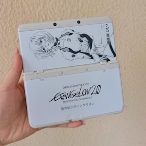 Puede incluir: Una consola de juegos portátil blanca con una ilustración en blanco y negro de un personaje en la parte superior. La sección inferior muestra el texto Groundwork of Evangelion: 2.0 You Can (Not) Advance y caracteres japoneses.