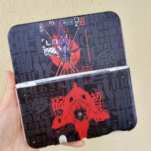 Op de afbeelding: Een zwarte draagbare spelcomputer met een grafisch ontwerp geïnspireerd op Neon Genesis Evangelion. De bovenklep toont een paarse mecha-figuur tegen een rode cirkel, terwijl de onderklep een grote rode abstracte vorm en Japanse tekst bevat.