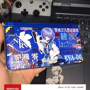 Könnte beinhalten: Eine blaue tragbare Spielkonsole mit einem Grafik-Skin von Rei Ayanami aus Neon Genesis Evangelion. Das Design umfasst das NERV-Logo, japanischen Text, den Schriftzug PROTOTYPE EVA und die Figur, die eine schwarze Handfeuerwaffe hält.
