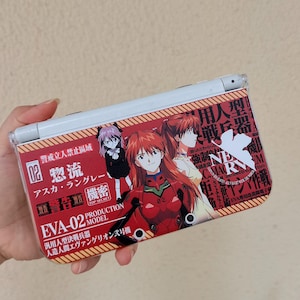 May include: A white handheld gaming console featuring a decorative skin with anime characters Asuka Langley and Rei Ayanami. The design includes red and orange diagonal stripes, Japanese text, the text EVA-02 PRODUCTION MODEL, and a white NERV logo.