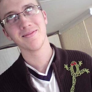 Op de afbeelding: Een jonge persoon met een bril en een bruine trui met een groene gehaakte kikker broche. De kikker heeft rode ogen en draagt ​​een strikje.