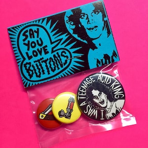 May include: A set of four round pinback buttons. The buttons are black, white, and yellow. The buttons feature a variety of designs, including a person with a crown, a person with a cigarette, and a person with a knife. The buttons are packaged in a clear plastic bag with a black and white card that says "Say You Love Buttons".