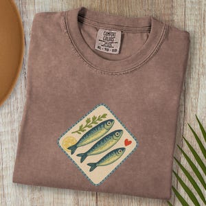 以下が含まれることがあります： エスプレッソ色のTシャツ、魚のグラフィック入り。グラフィックは、3匹の青い魚、レモンスライス、葉の小枝、赤いハートが特徴です。シャツはクルーネックで、Comfort Colorsのタグが付いています。