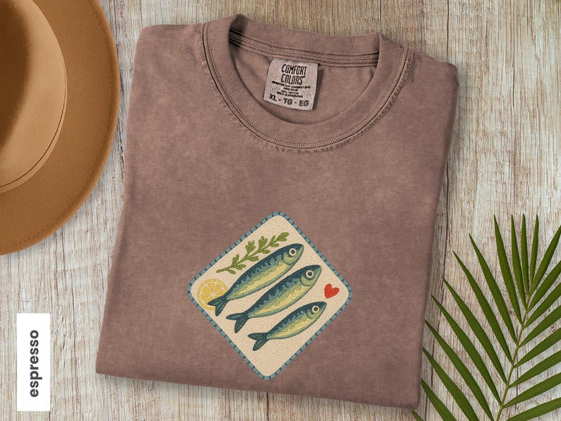 以下が含まれることがあります： エスプレッソ色のTシャツ、魚のグラフィック入り。グラフィックは、3匹の青い魚、レモンスライス、葉の小枝、赤いハートが特徴です。シャツはクルーネックで、Comfort Colorsのタグが付いています。