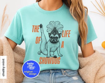 May include: A light blue t-shirt with the text "THE LIFE OF A SHOWDOG" in orange. The shirt features a drawing of a French bulldog wearing a feathered headdress. The shirt has a round, crew neck and short sleeves.