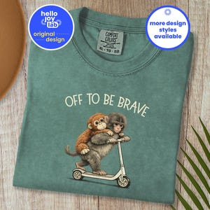 Op de afbeelding: Lichtgroen T-shirt met de tekst "OFF TO BE BRAVE" en een afbeelding van twee apen op een scooter. Het shirt heeft een ronde hals en korte mouwen. Het ontwerp bevat de tekst "hello joy lab original design" en "more design styles available".