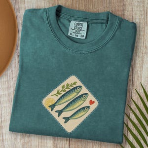 以下が含まれることがあります： 魚のグラフィックが施されたブルー・スプルースのTシャツ。グラフィックには、3匹の魚、レモンスライス、葉の小枝、赤いハートが描かれています。シャツのサイズはXL-TG-EGです。