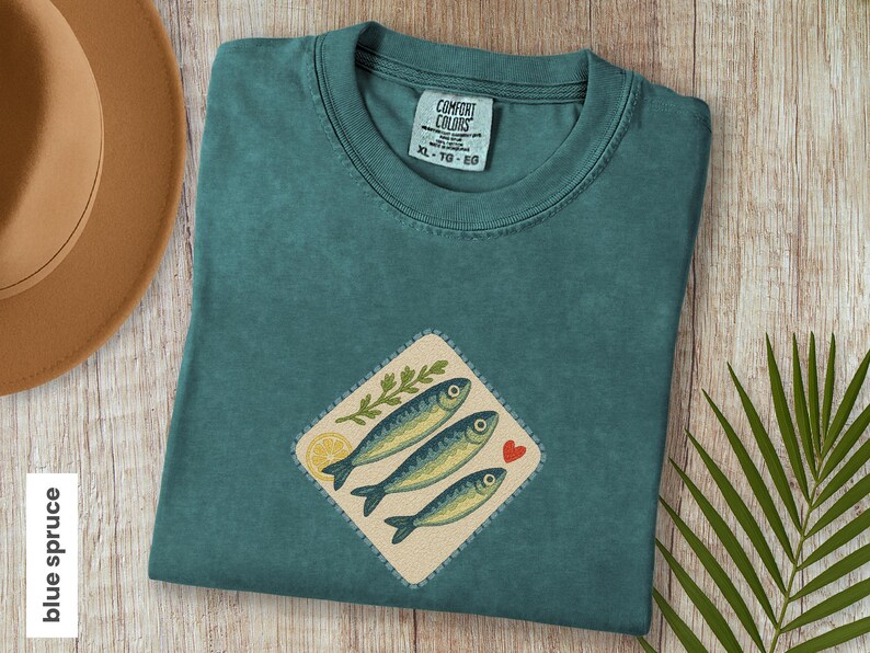 以下が含まれることがあります： 魚のグラフィックが施されたブルー・スプルースのTシャツ。グラフィックには、3匹の魚、レモンスライス、葉の小枝、赤いハートが描かれています。シャツのサイズはXL-TG-EGです。