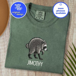 May include: A moss green crew neck t-shirt featuring a graphic illustration of a raccoon in the center. Below the raccoon, the name JIMOTHY is printed in white capital letters. The shirt has a Comfort Colors brand label at the neckline.