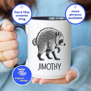May include: A white ceramic mug with a black interior and handle, featuring a black and white illustration of a raccoon and the text JIMOTHY printed below it. The mug includes text labels stating 11oz & 15oz ceramic mug and more phrases available.