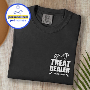K&ouml;nnte beinhalten: Schwarzes T-Shirt mit dem Text &bdquo;TREAT DEALER&ldquo; und zwei Knochen-Grafiken. Das Shirt zeigt auch die Namen &bdquo;ZAHRA &bull; RUBY&ldquo; und eine Grafik eines Hundekopfes. Ein kreisf&ouml;rmiges Grafikfeld zeigt &bdquo;personalisierte Haustiernamen&ldquo;.