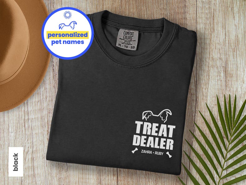 K&ouml;nnte beinhalten: Schwarzes T-Shirt mit dem Text &bdquo;TREAT DEALER&ldquo; und zwei Knochen-Grafiken. Das Shirt zeigt auch die Namen &bdquo;ZAHRA &bull; RUBY&ldquo; und eine Grafik eines Hundekopfes. Ein kreisf&ouml;rmiges Grafikfeld zeigt &bdquo;personalisierte Haustiernamen&ldquo;.