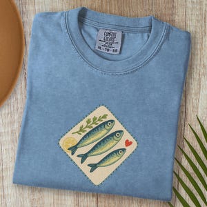 以下が含まれることがあります： 魚のデザインが施された水色のTシャツ。デザインには、3匹の魚、レモンスライス、葉の小枝、赤いハートが含まれています。シャツにはXL-TG-EGと表示されています。
