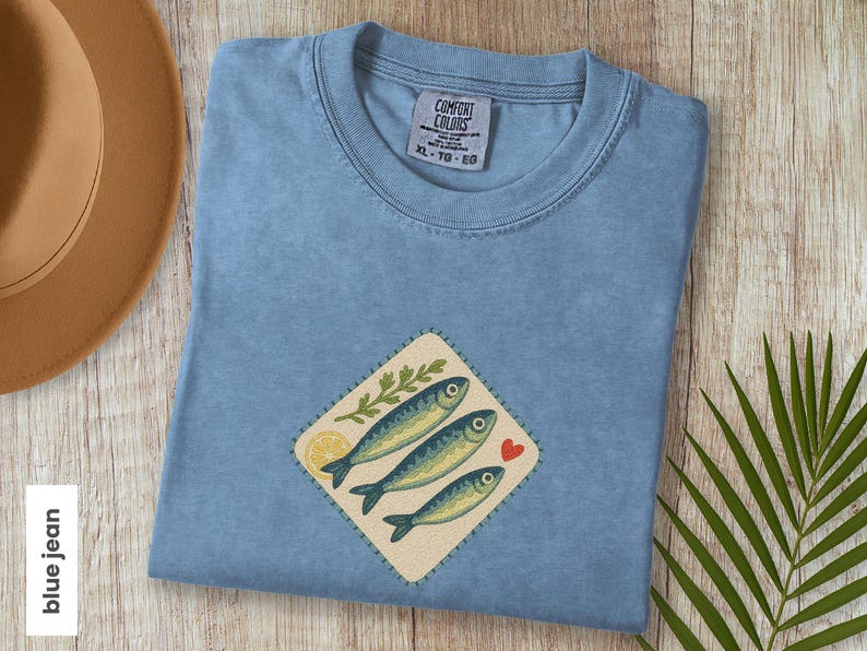 以下が含まれることがあります： 魚のデザインが施された水色のTシャツ。デザインには、3匹の魚、レモンスライス、葉の小枝、赤いハートが含まれています。シャツにはXL-TG-EGと表示されています。