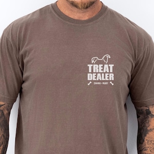 K&ouml;nnte beinhalten: Braunes T-Shirt mit dem Text "TREAT DEALER" und einer Hunde-Silhouette. Darunter stehen die Namen "ZAHRA &bull; RUBY" und Knochen-Grafiken. Das Shirt hat kurze &Auml;rmel und einen Rundhalsausschnitt.