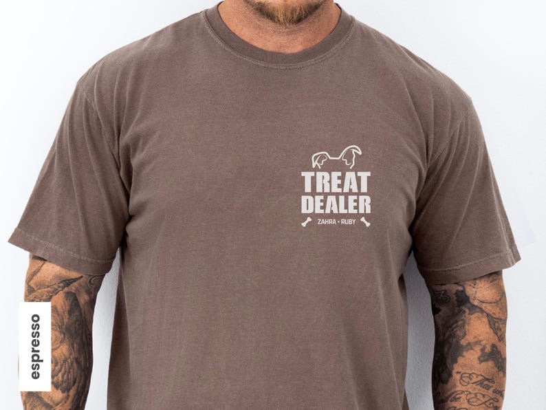 K&ouml;nnte beinhalten: Braunes T-Shirt mit dem Text "TREAT DEALER" und einer Hunde-Silhouette. Darunter stehen die Namen "ZAHRA &bull; RUBY" und Knochen-Grafiken. Das Shirt hat kurze &Auml;rmel und einen Rundhalsausschnitt.