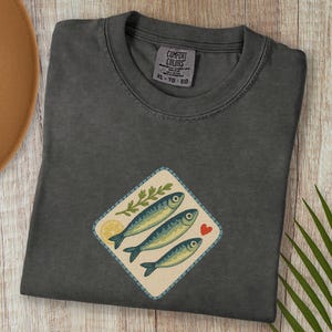 以下が含まれることがあります： 3匹の青い魚、レモンスライス、緑の小枝、赤いハートが描かれたひし形のパッチが付いた、ダークグレーのComfort Colors Tシャツ。木製の表面に折りたたまれており、帽子とヤシの葉が見えます。