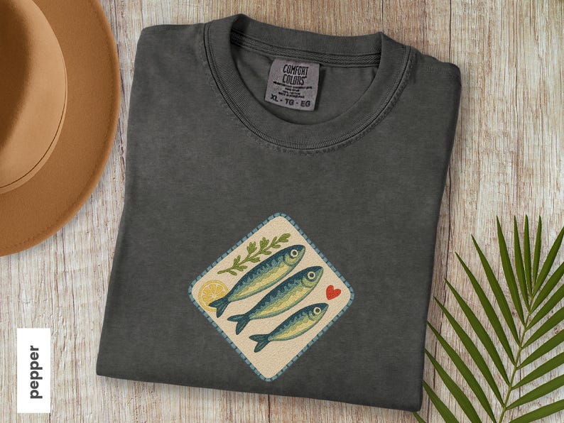 以下が含まれることがあります： 3匹の青い魚、レモンスライス、緑の小枝、赤いハートが描かれたひし形のパッチが付いた、ダークグレーのComfort Colors Tシャツ。木製の表面に折りたたまれており、帽子とヤシの葉が見えます。