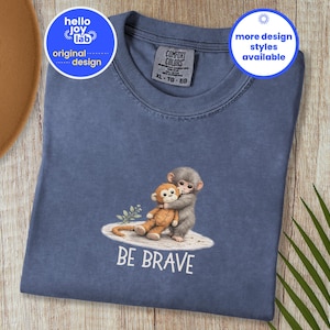 Könnte beinhalten: Denimblaues T-Shirt mit einer Grafik von zwei sich umarmenden Affen und dem Text "BE BRAVE" in Weiß. Das T-Shirt hat einen Rundhalsausschnitt und ist aus weichem Material. Das Shirt ist von der Marke Comfort Colors, Größe XL-TG.