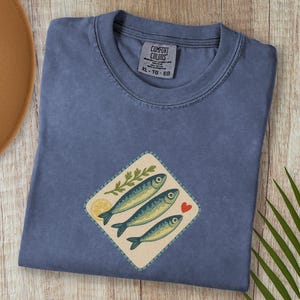 以下が含まれることがあります： デニムブルーのTシャツ。魚のグラフィックが特徴です。グラフィックには3匹の魚、レモンスライス、緑の小枝、赤いハートが含まれています。シャツのサイズはXLです。タグには「Comfort Colors」と書かれています。