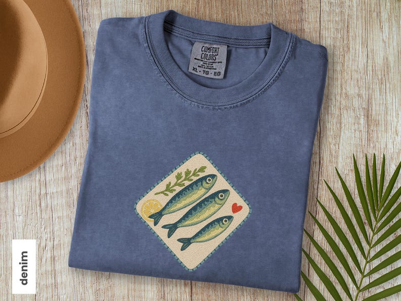以下が含まれることがあります： デニムブルーのTシャツ。魚のグラフィックが特徴です。グラフィックには3匹の魚、レモンスライス、緑の小枝、赤いハートが含まれています。シャツのサイズはXLです。タグには「Comfort Colors」と書かれています。
