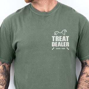 K&ouml;nnte beinhalten: Moosgr&uuml;nes T-Shirt mit dem Text "TREAT DEALER" und einer Hunde-Silhouette. Darunter stehen die Namen "ZAHRA &bull; RUBY" und zwei Knochen-Grafiken. Das Shirt hat eine einfarbige Farbe.