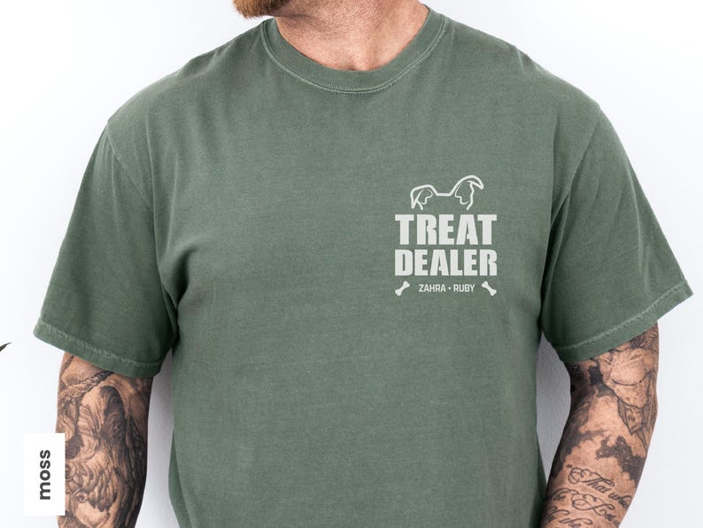 K&ouml;nnte beinhalten: Moosgr&uuml;nes T-Shirt mit dem Text "TREAT DEALER" und einer Hunde-Silhouette. Darunter stehen die Namen "ZAHRA &bull; RUBY" und zwei Knochen-Grafiken. Das Shirt hat eine einfarbige Farbe.
